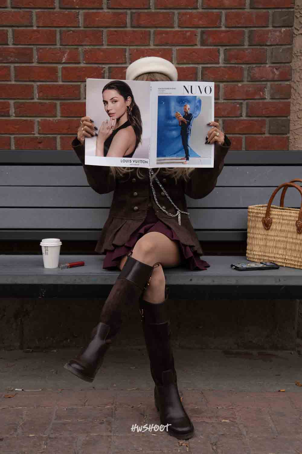 Seated on a grey bench, Raina holds an open NUVO magazine in front of her face, with coffee and a basket bag beside her.