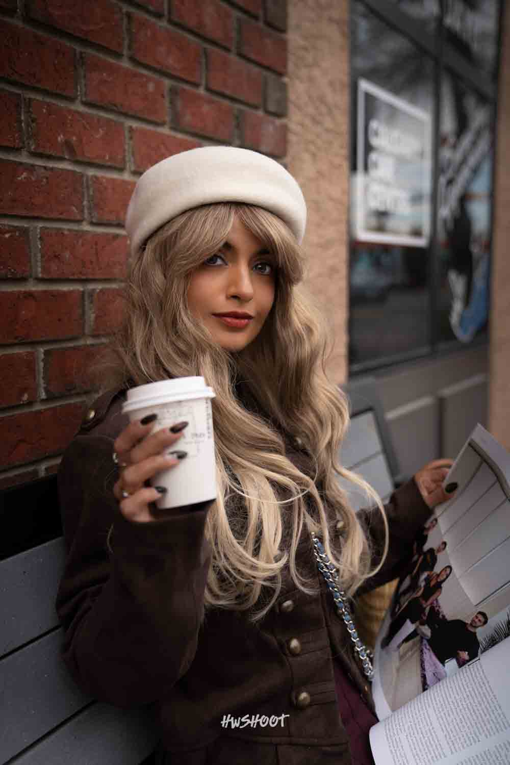 Raina looks toward the camera while holding a takeaway cup and magazine beside an Inglewood brick wall.