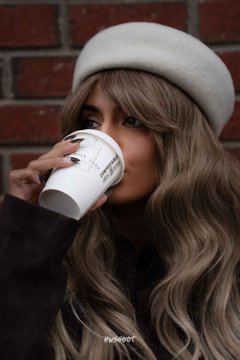 Close-up of Raina sipping coffee, with a white beret, long blonde waves and a red brick background.