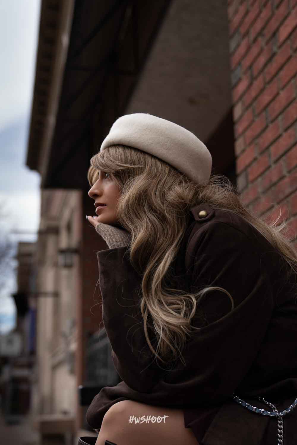 Raina rests her chin on her hand in profile beside a brick storefront, wearing a brown jacket and white beret.