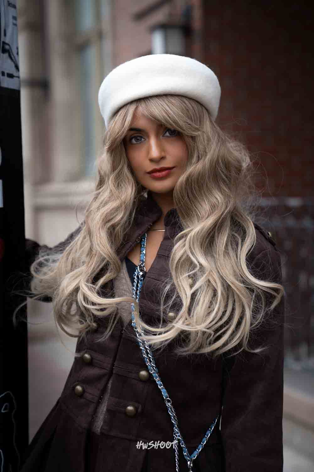 Raina faces the camera in a double-breasted brown jacket and white beret, with a silver chain across her outfit.