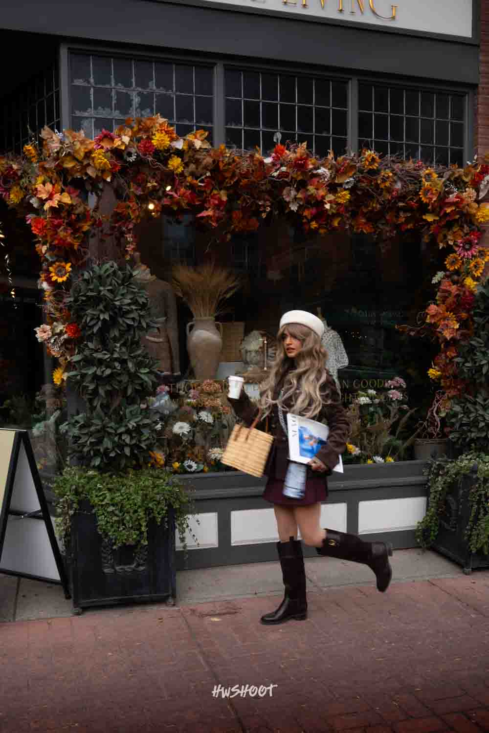 Raina lifts one boot behind her outside a flower-framed shop window, carrying coffee, a magazine and a woven bag.